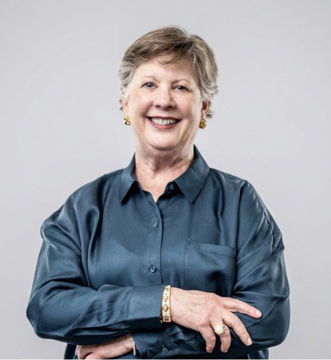 Smiling older woman with short gray hair, wearing a teal satin blouse and gold jewelry, arms crossed lightly in front of a light gray background.